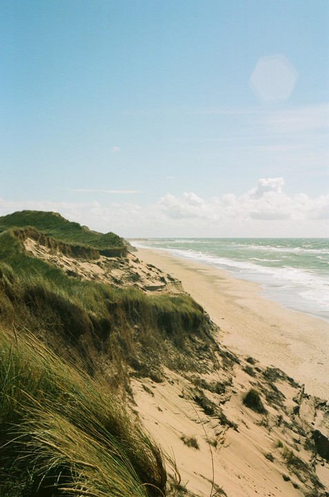 A sandy beach with green dunes and ocean waves under a blue sky.