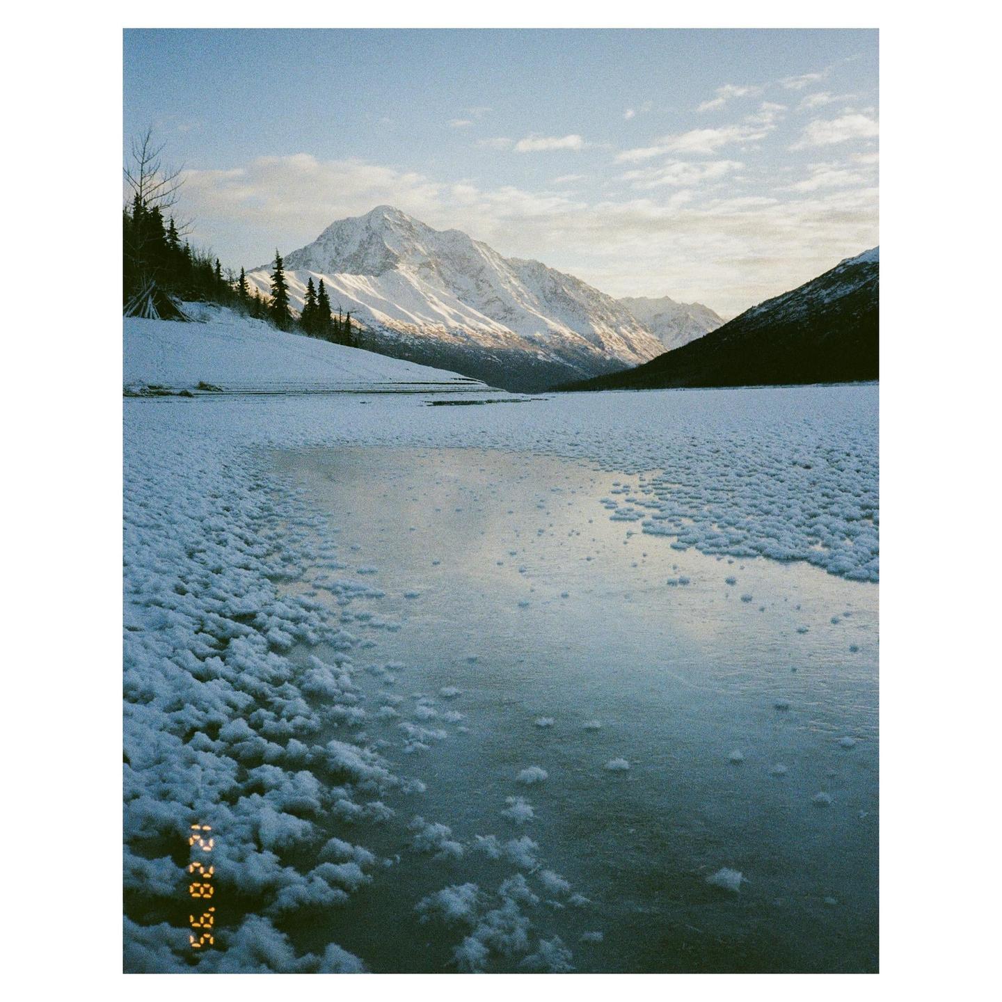 A frozen lake with snow-covered banks is surrounded by mountains under a cloudy sky.