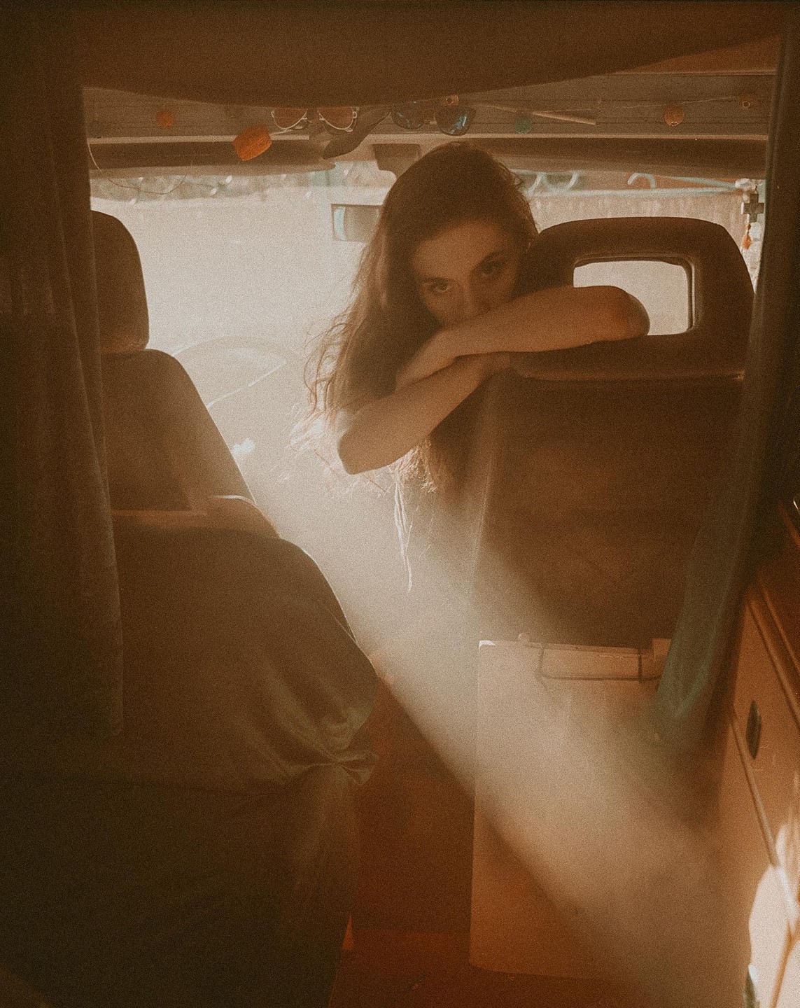 A woman with long hair leans on a car seat, looking directly at the viewer as a ray of light illuminates the interior of the vehicle.
