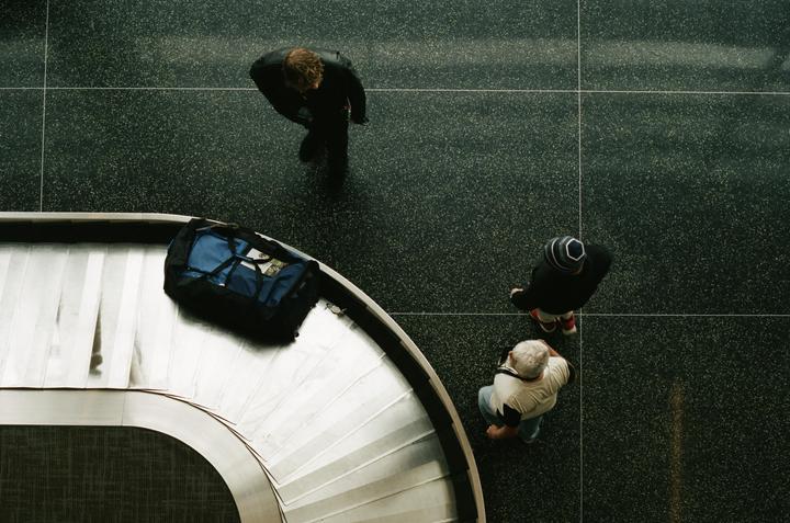 Three people stand around a baggage claim carousel with a blue duffel bag.