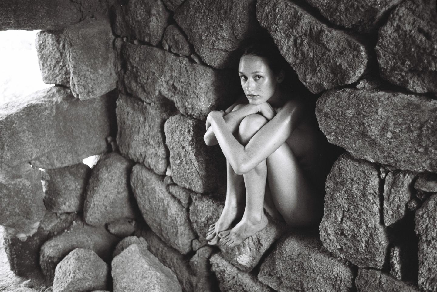 A woman sits curled up in a stone ruin, looking towards the viewer.