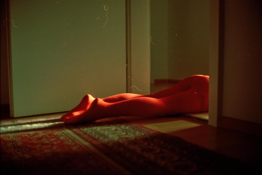 A person's legs are visible on a rug in a dimly lit room, with light casting shadows on the floor.