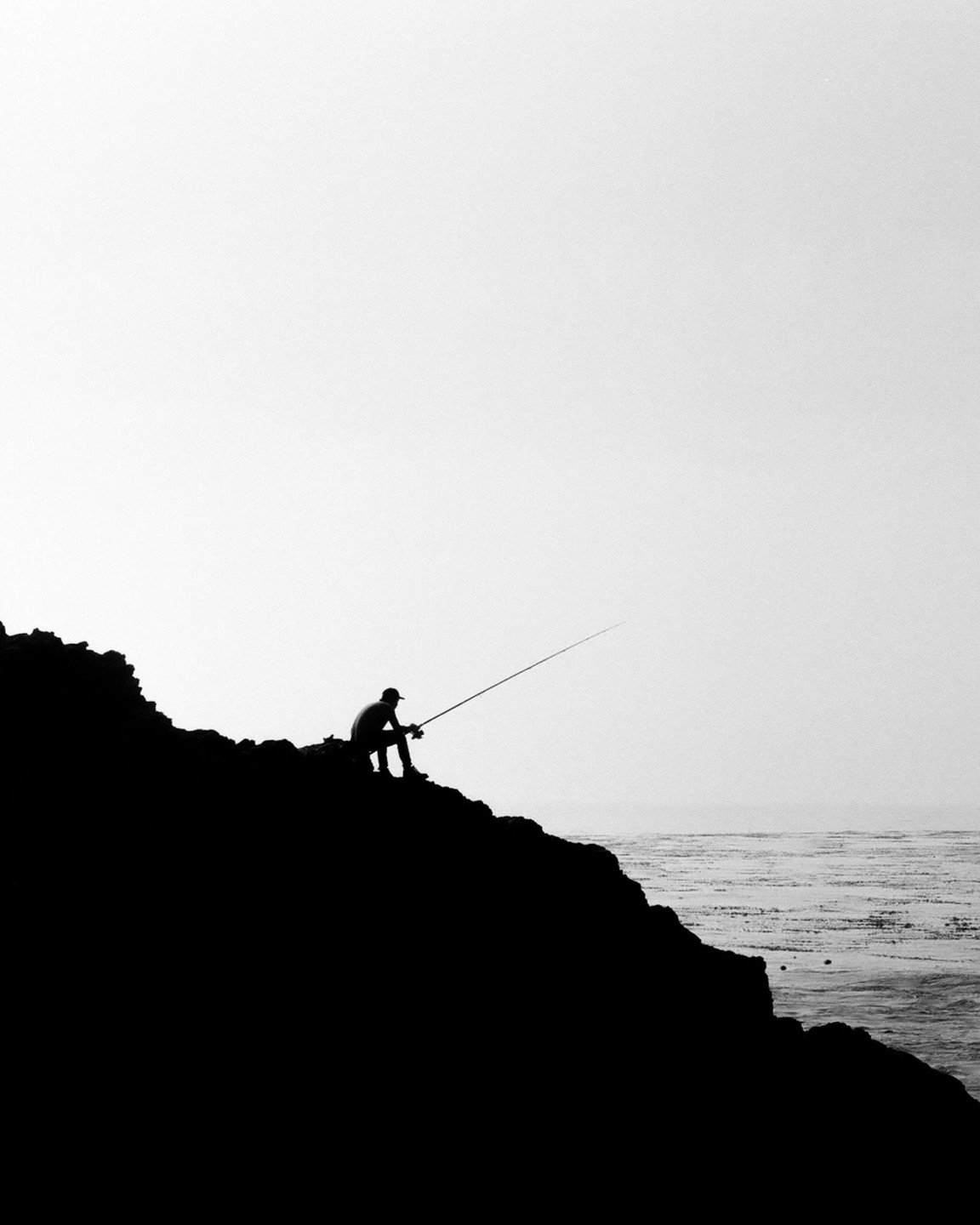 A man sits on a cliff fishing into the ocean.