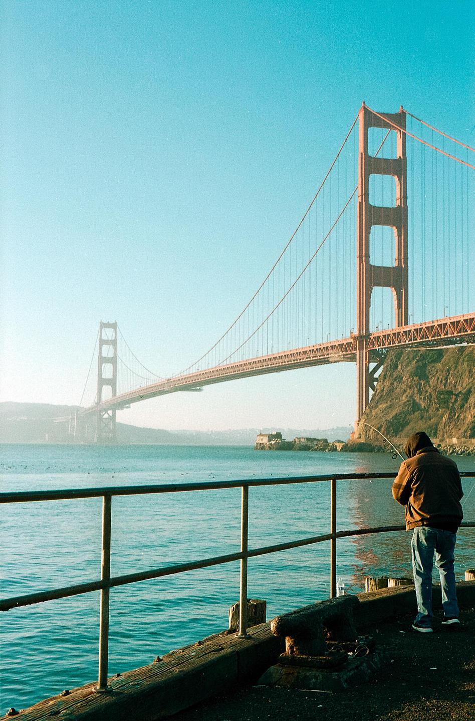 A man fishes from a pier with the Golden Gate Bridge in the background.