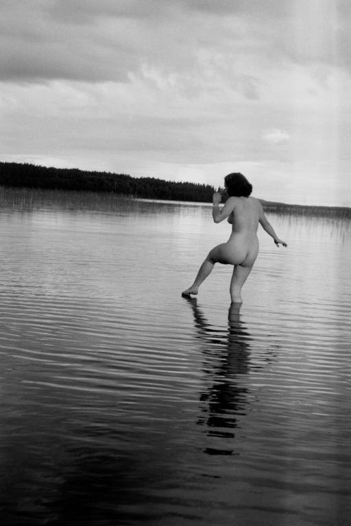 A nude woman stands in a lake with a forest in the background under a cloudy sky.