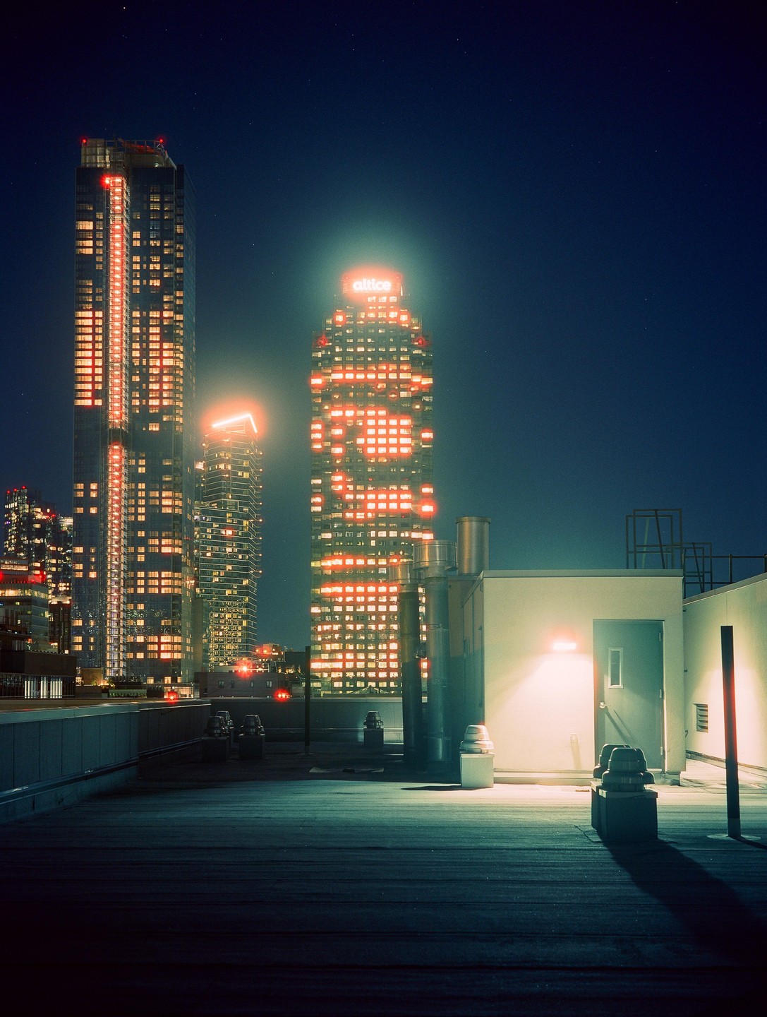 A rooftop with industrial elements overlooks a city skyline at night, with illuminated skyscrapers in the background.