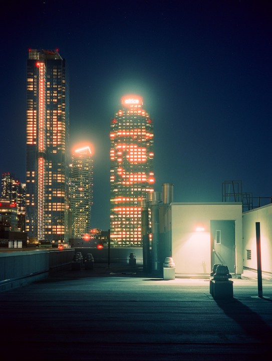 A rooftop with industrial elements overlooks a city skyline at night, with illuminated skyscrapers in the background.