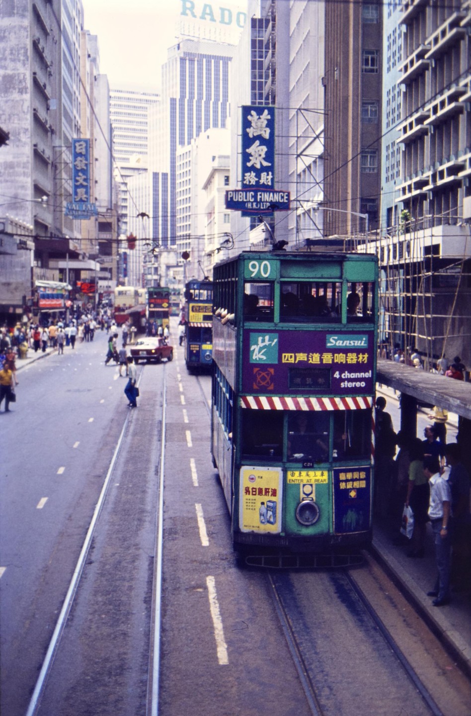A green double-decker tram is stopped at a station on a busy street in Hong Kong.