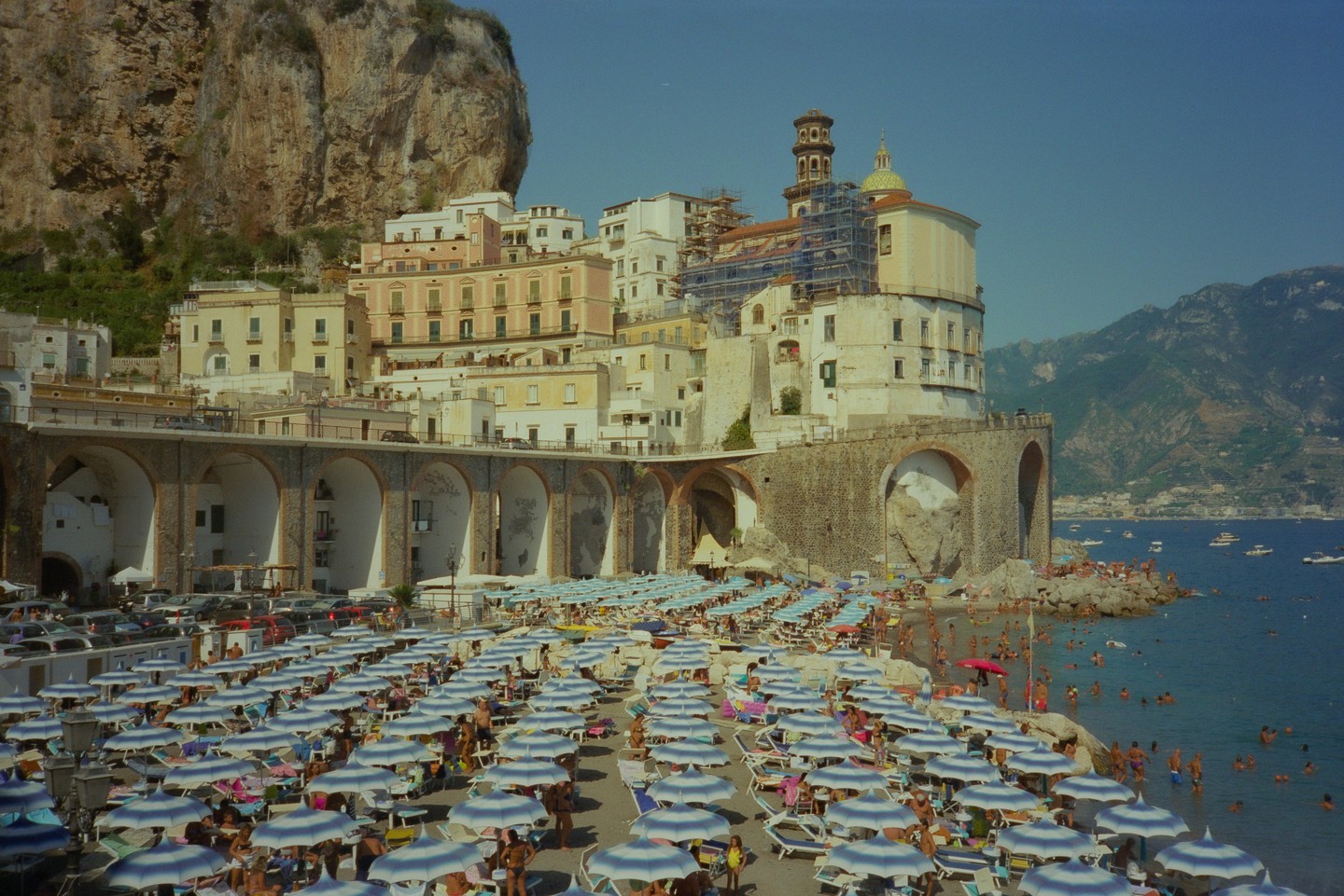 A crowded beach with many umbrellas is next to a town built into a cliff face overlooking the sea.