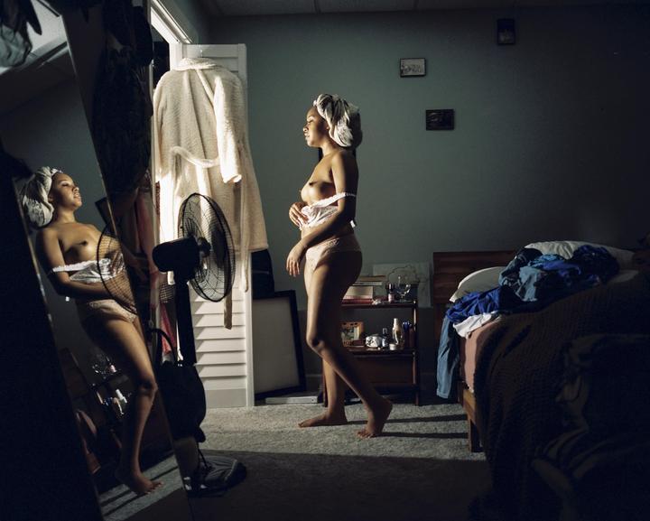 A woman stands in a bedroom, partially dressed, with her reflection visible in a mirror.