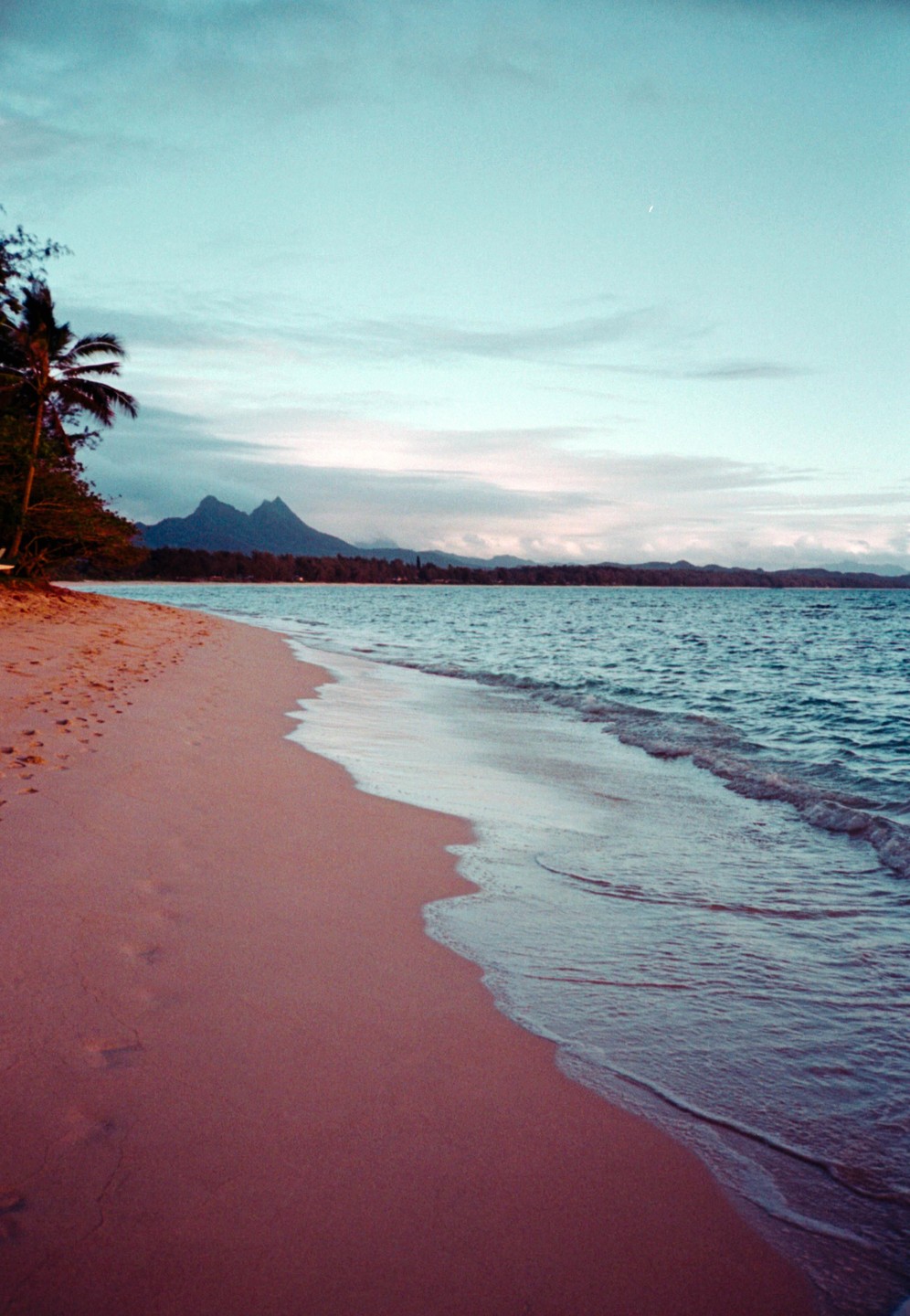 A sandy beach with gentle waves meets a distant mountain range under a cloudy sky at sunrise.