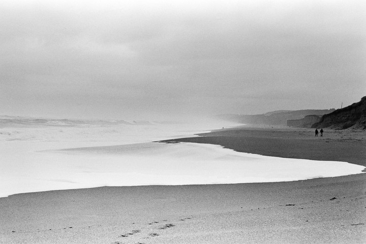Two people walk along a wide, sandy beach with waves crashing on the shore and cliffs in the distance.