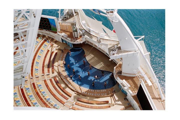 A high-angle view shows people in a pool on a cruise ship deck with rows of empty lounge chairs and the ocean in the background.