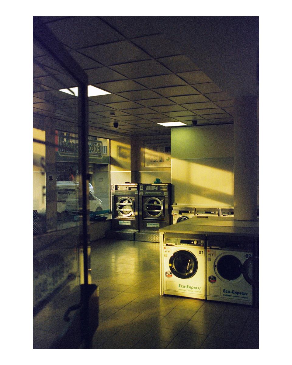Sunlight streams into a laundromat with several washing machines and dryers.