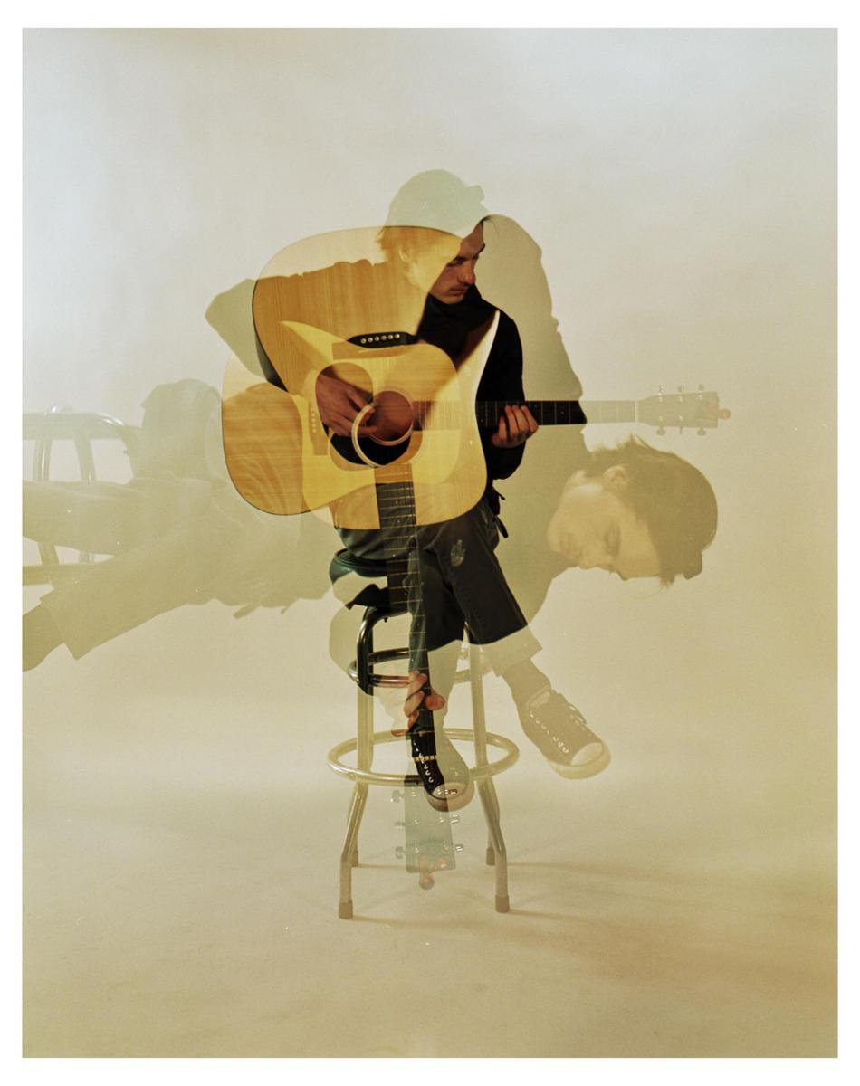 A man sits on a stool playing an acoustic guitar, with multiple exposures creating a ghostly effect.
