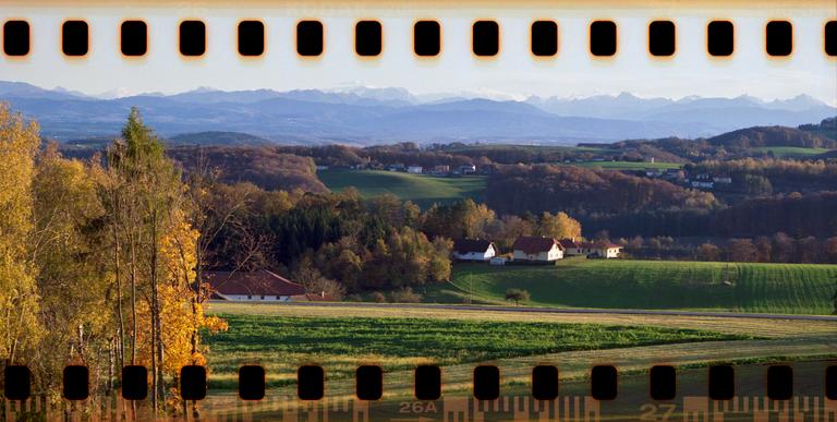 A rural landscape with houses and fields in the foreground, autumn trees in the middle ground, and mountains in the distance.