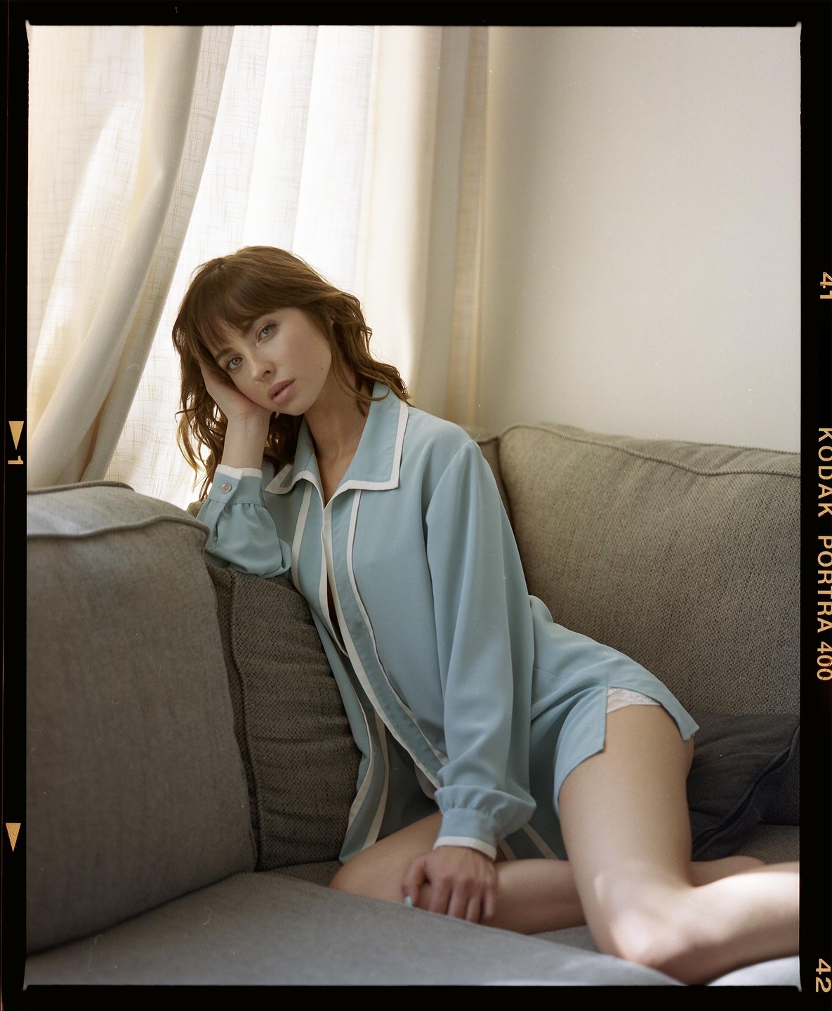 A woman in a light blue pajama set is sitting on a gray sofa next to a window with sheer curtains.