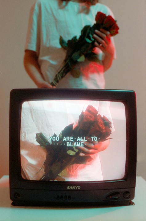 A television displays a person holding a bouquet of roses with the text "YOU ARE ALL TO BLAME" superimposed.