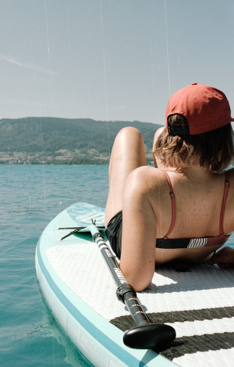 A woman in a bikini and red cap is lying on a paddleboard in a lake with mountains in the background.