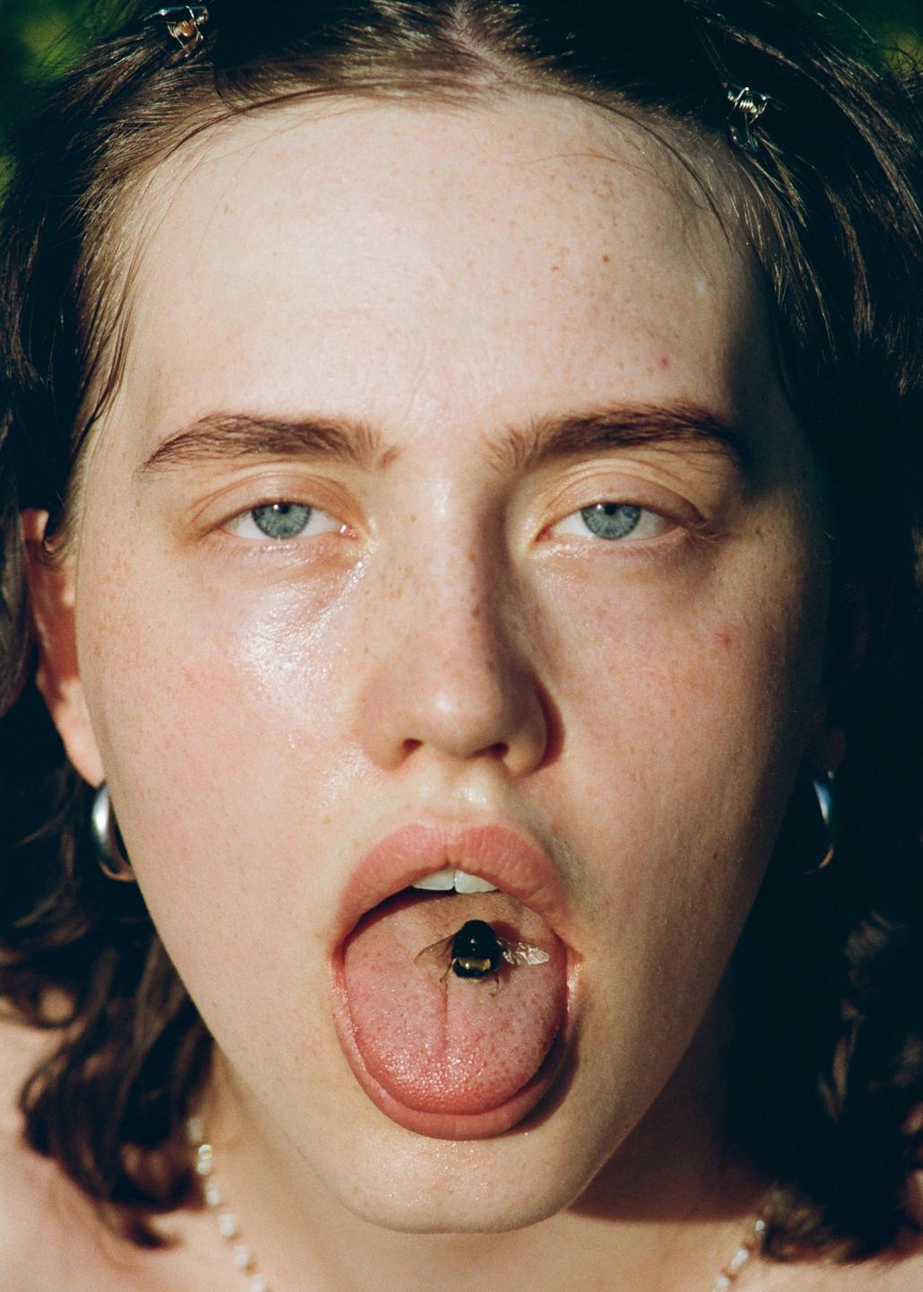 A woman with a bumblebee on her tongue stares directly at the viewer.