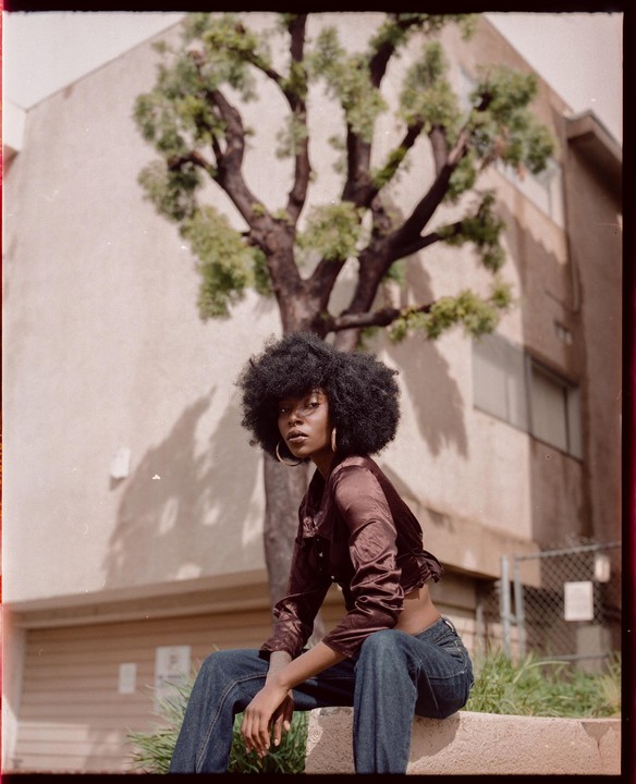 A woman with an afro hairstyle and hoop earrings sits outdoors in front of a building and a tree.