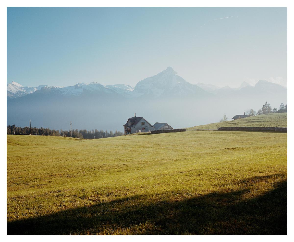 A house sits on a hill overlooking a green field with mountains in the background.