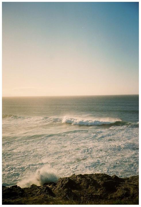 A large wave breaks on the shore of a rocky coastline under a clear sky.