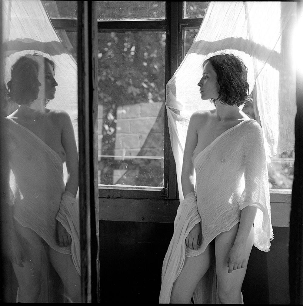 A woman draped in sheer fabric stands in front of a window, looking to the side, with her reflection visible in a mirror.
