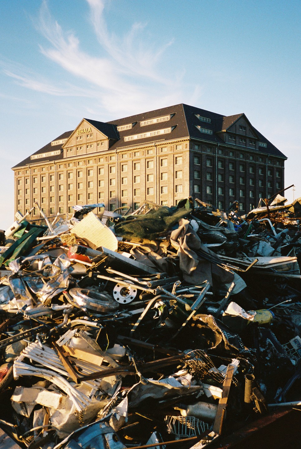 A large pile of scrap metal sits in front of a grand brick building under a blue sky.