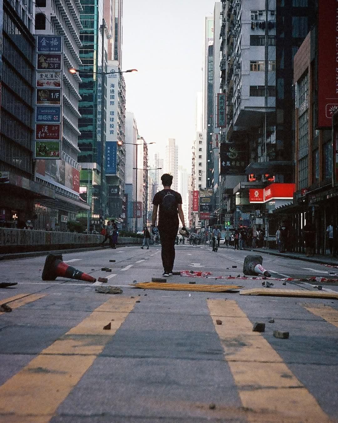 A man walks down a street littered with debris and traffic cones, surrounded by tall buildings with signs.