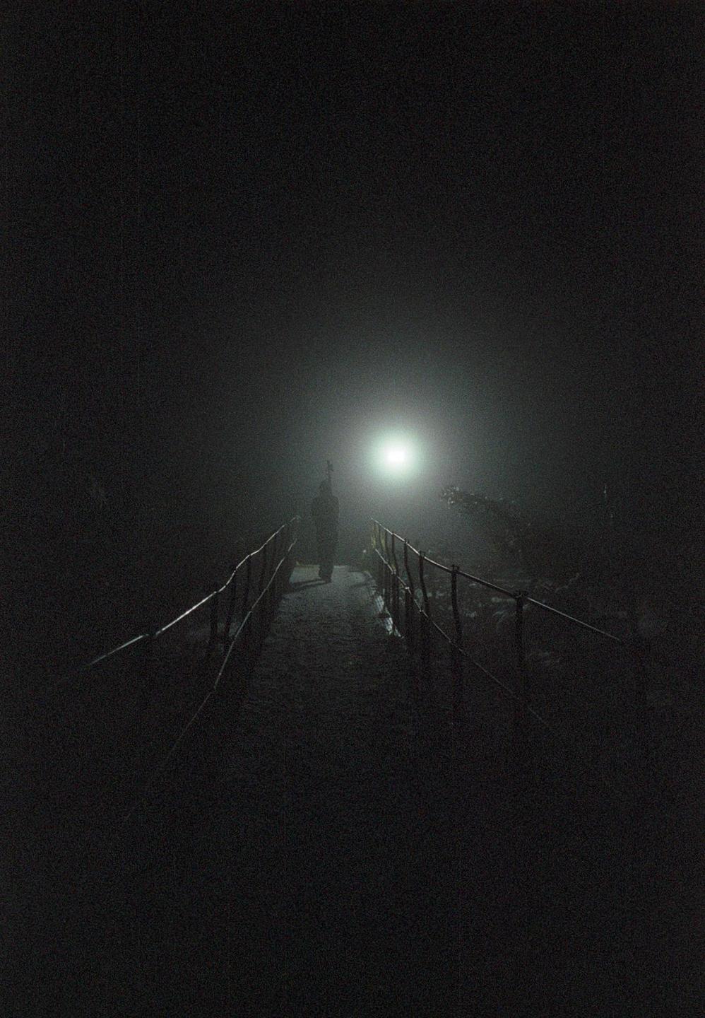 A person walks on a narrow path with railings on both sides, illuminated by a bright light in the distance.