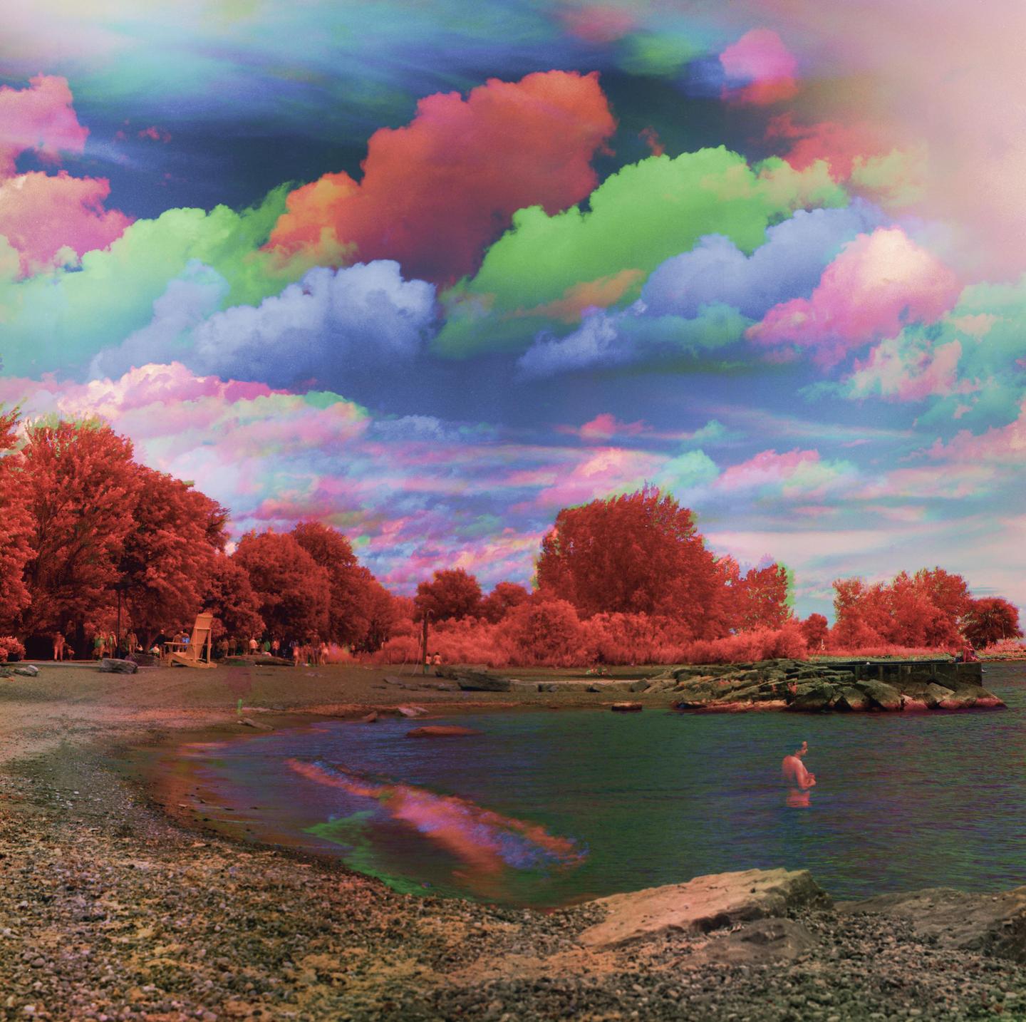 A man stands in the water at a rocky beach with colorful clouds in the sky.