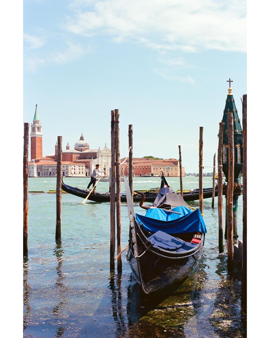 A gondola is moored to wooden posts in the foreground, with a gondolier standing in another gondola in the background, and a church and other buildings across the water.