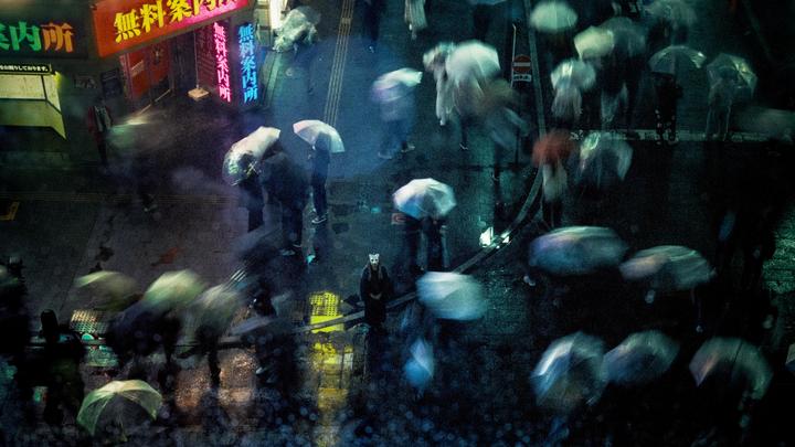 A person in a cat mask stands in the middle of a rainy street at night, surrounded by blurred figures holding umbrellas and neon signs.