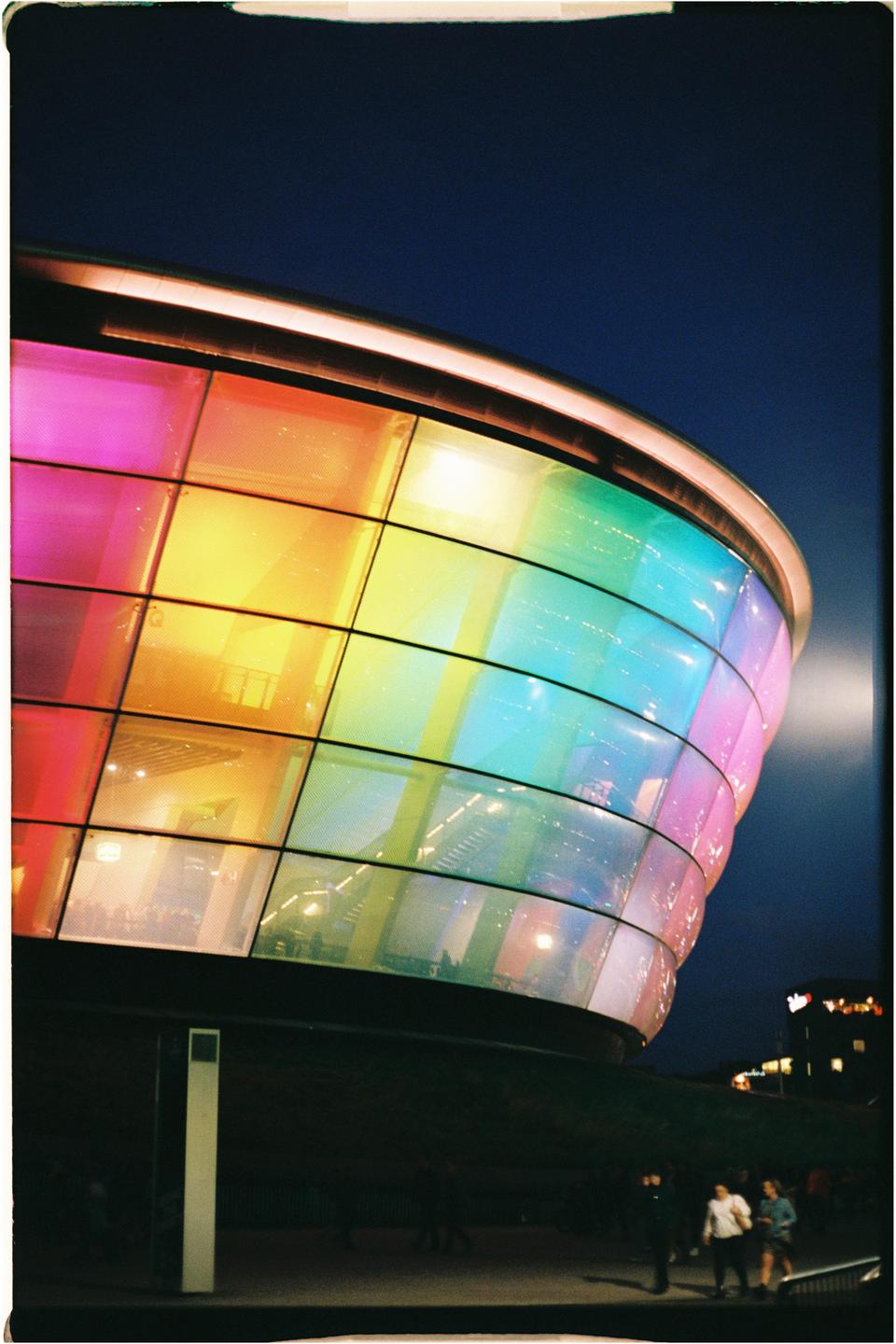 A building with a curved, rainbow-colored glass facade stands against a dark blue night sky.