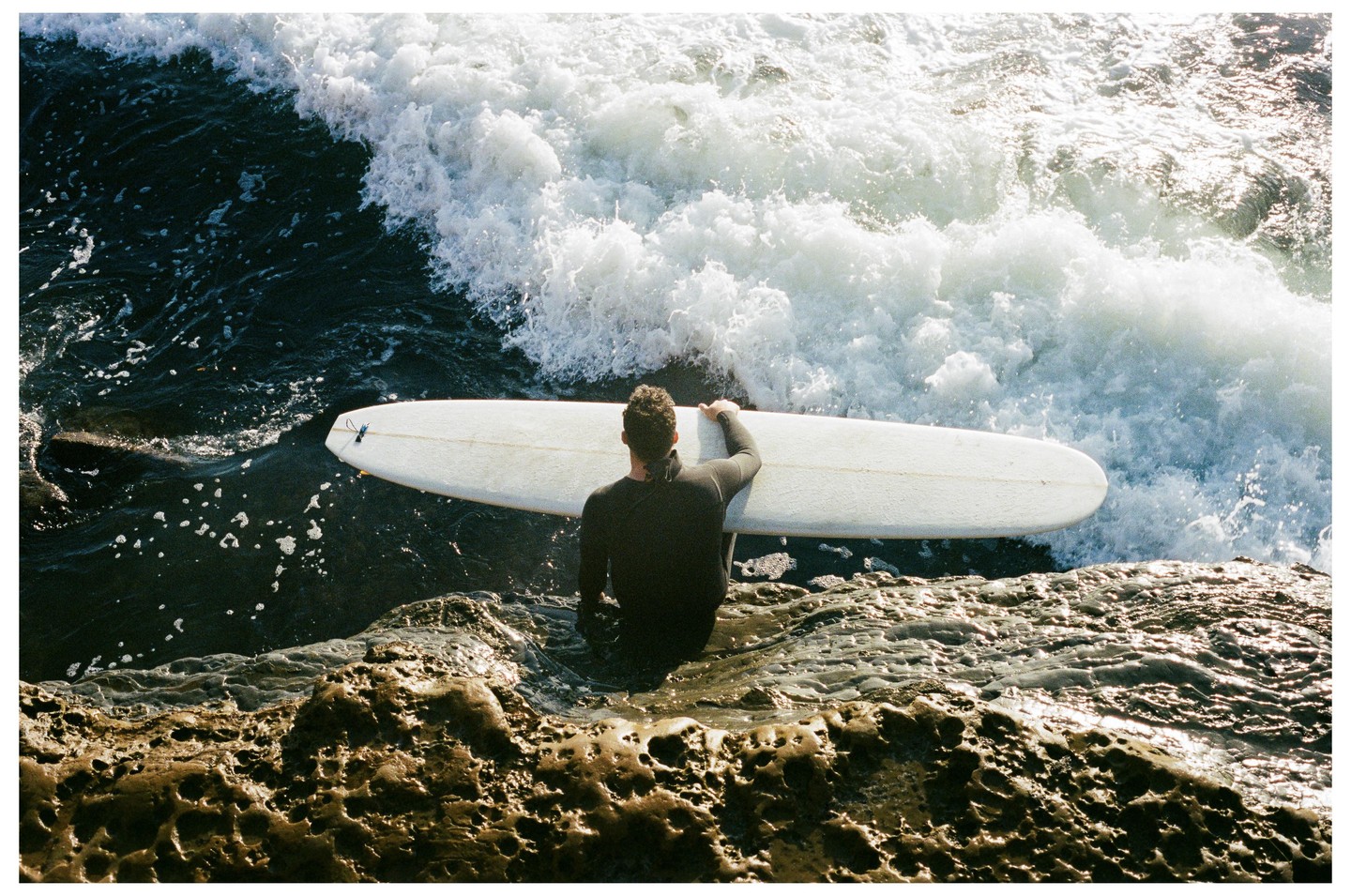 A surfer in a wetsuit holds a surfboard in the water next to a rocky shore as a wave breaks.