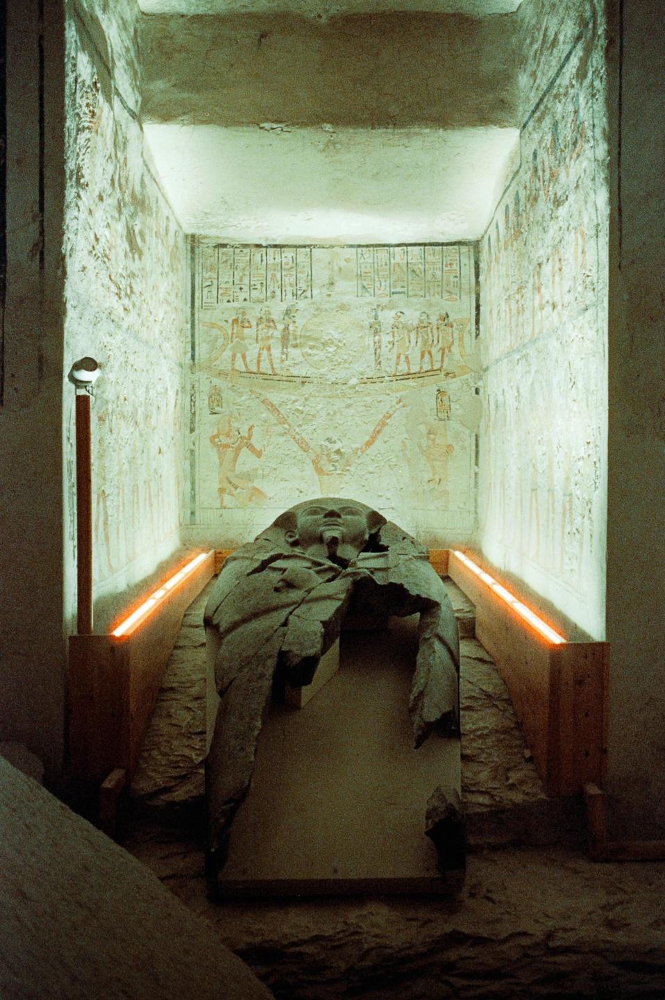 A stone sarcophagus is illuminated by orange lights in a tomb with painted hieroglyphs on the walls.