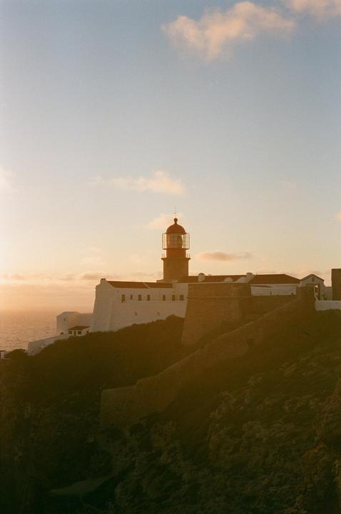 A lighthouse and white buildings are visible on a cliff overlooking the ocean at sunset.