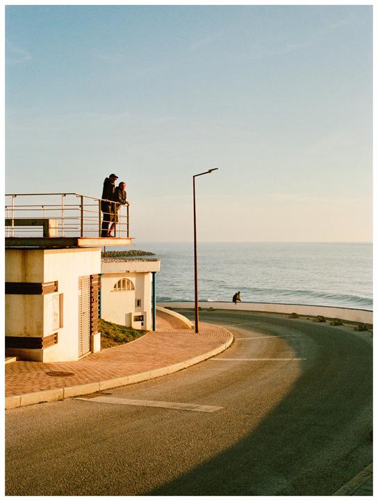 A couple stands on a balcony overlooking a coastal road and the ocean during golden hour.