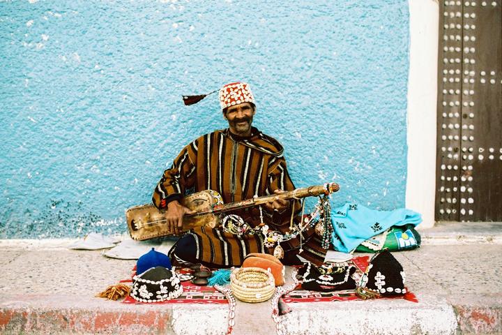 A man in traditional clothing sits on a patterned rug, playing a stringed instrument and surrounded by various crafts against a blue wall.