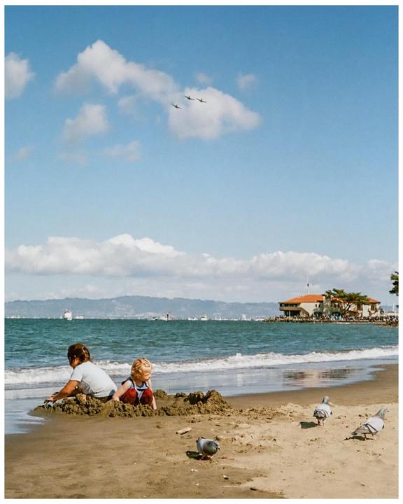 Two children play in the sand on a beach with pigeons nearby, while three airplanes fly in formation overhead.