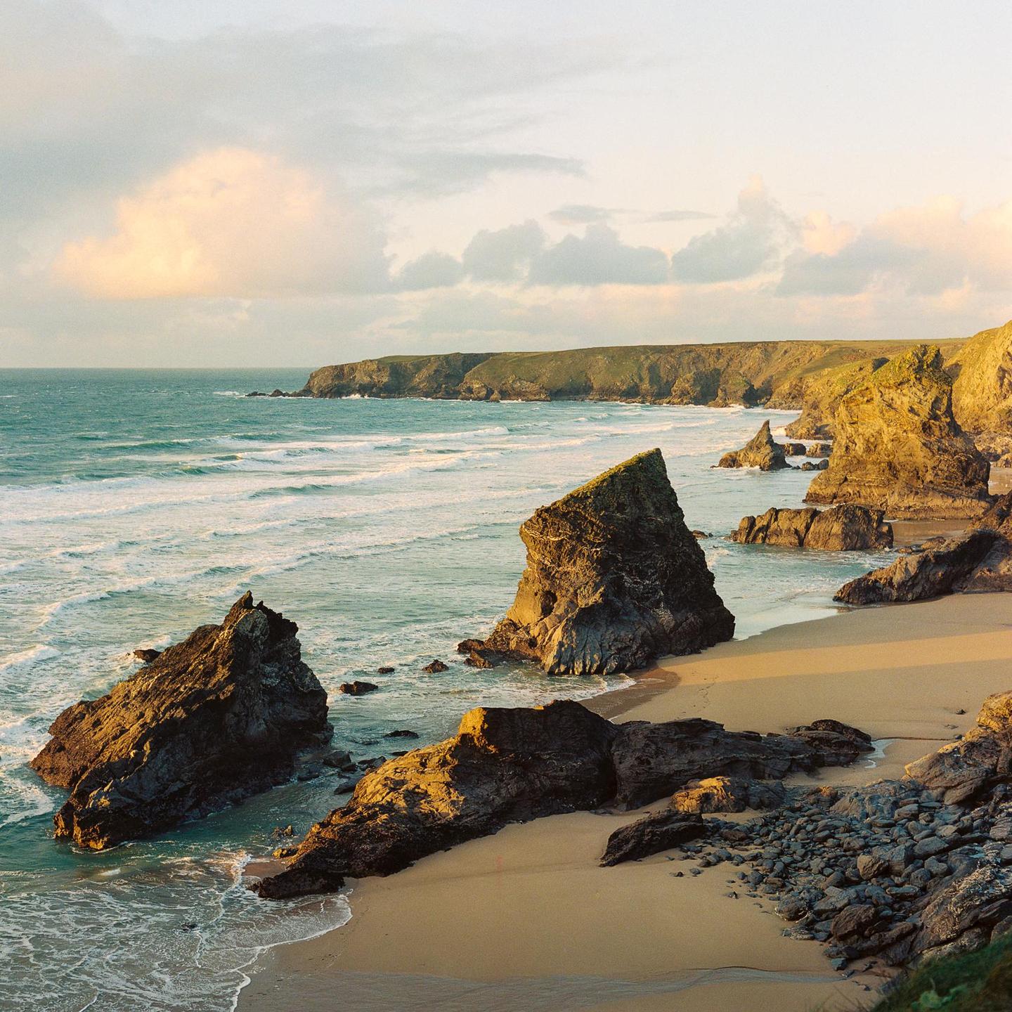A sandy beach with large rock formations is visible next to a cliff with green grass on top, with waves crashing on the shore.