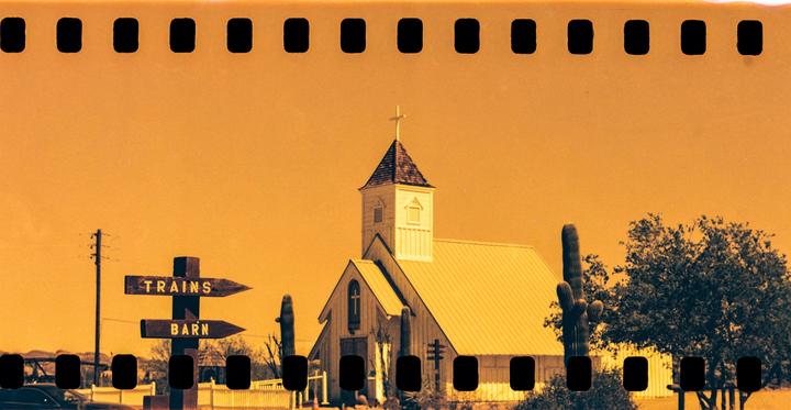 A church with a steeple and cross stands next to a saguaro cactus and a wooden sign pointing to "TRAINS" and "BARN" under an orange sky.