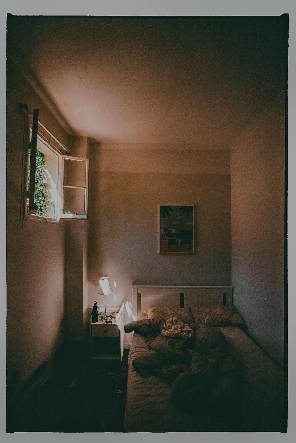 Sunlight streams through an open window into a bedroom with an unmade bed and a bedside table with a lamp.