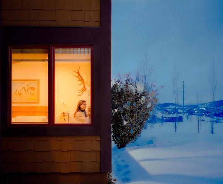 A woman looks out a window from a warmly lit room into a snowy landscape at dusk.