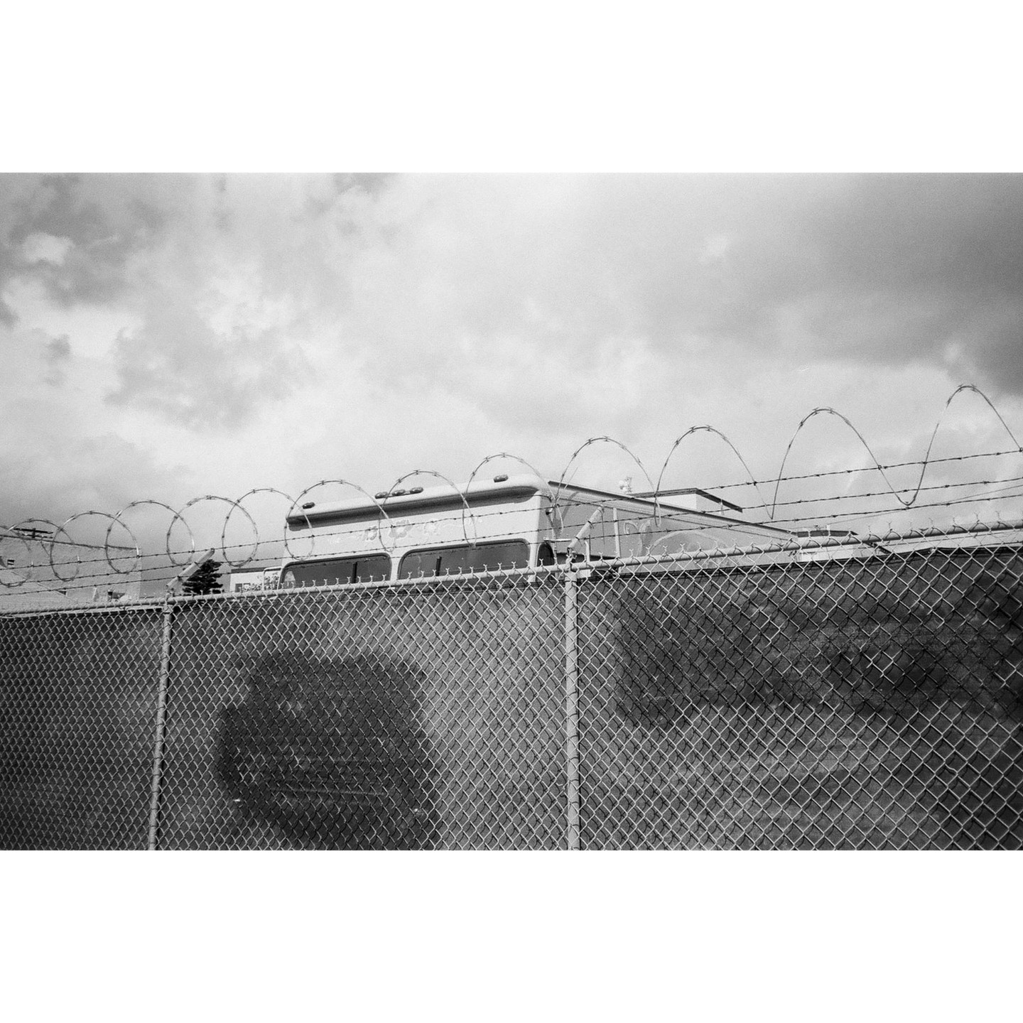 A bus is visible behind a chain link fence topped with razor wire under a cloudy sky.