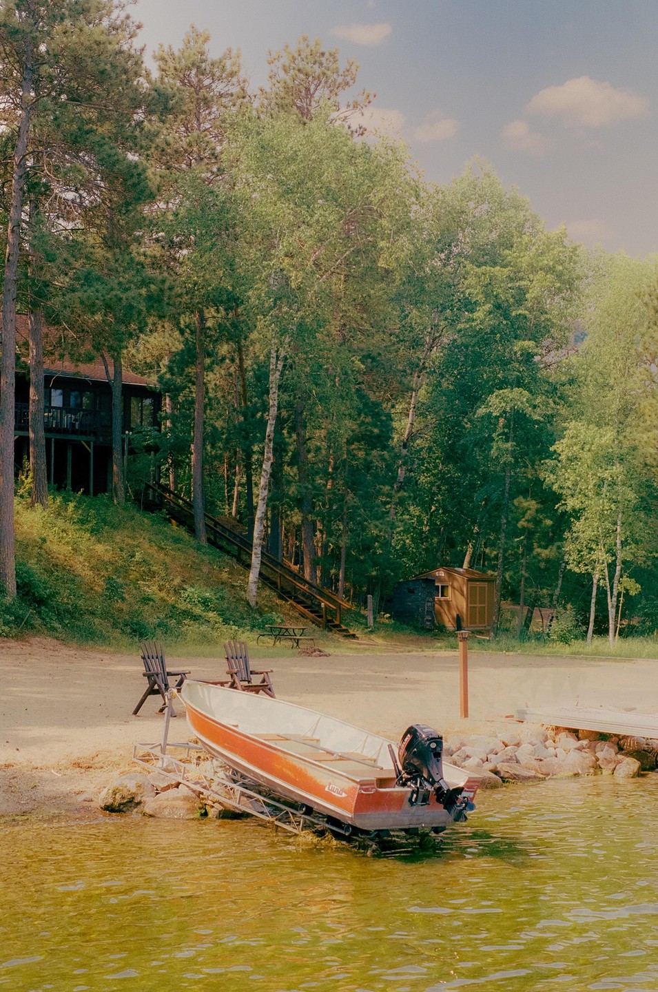 A boat is parked on a sandy shore next to a body of water, with a house and trees in the background.
