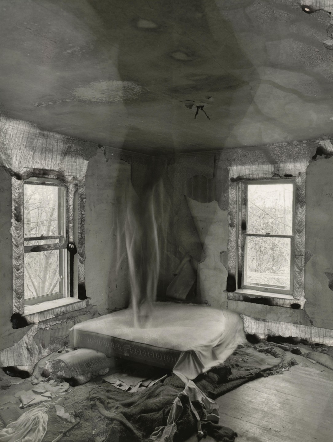 A ghostly figure hovers above a mattress in a dilapidated room with two windows.
