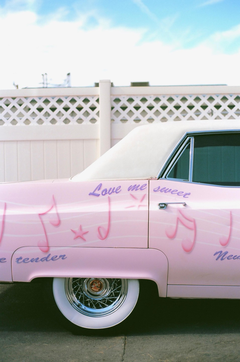A pink car with musical notes and text painted on its side is parked next to a white fence.