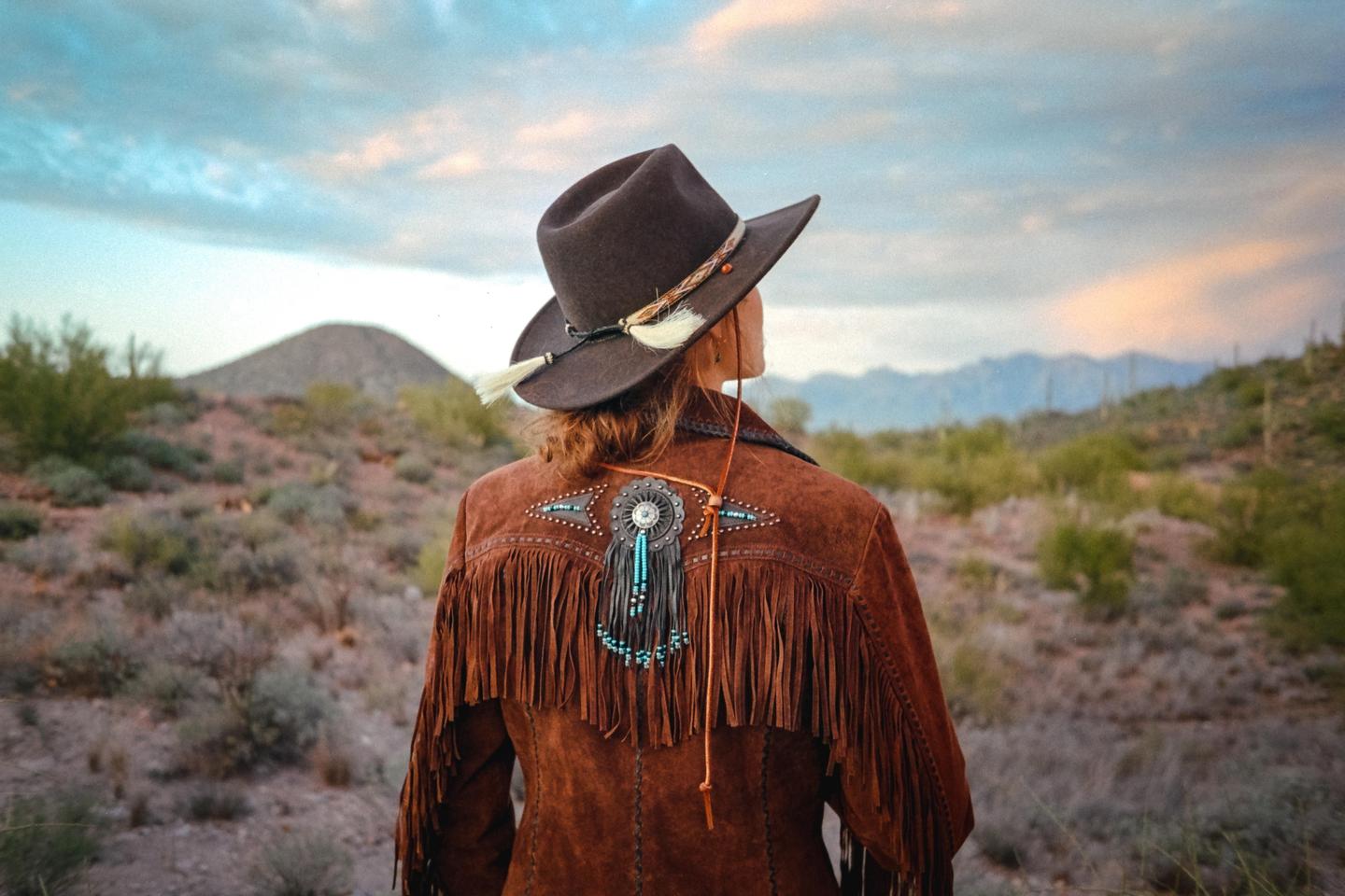 A woman wearing a cowboy hat and a fringed jacket stands with her back to the viewer, looking out at a desert landscape with mountains in the distance.